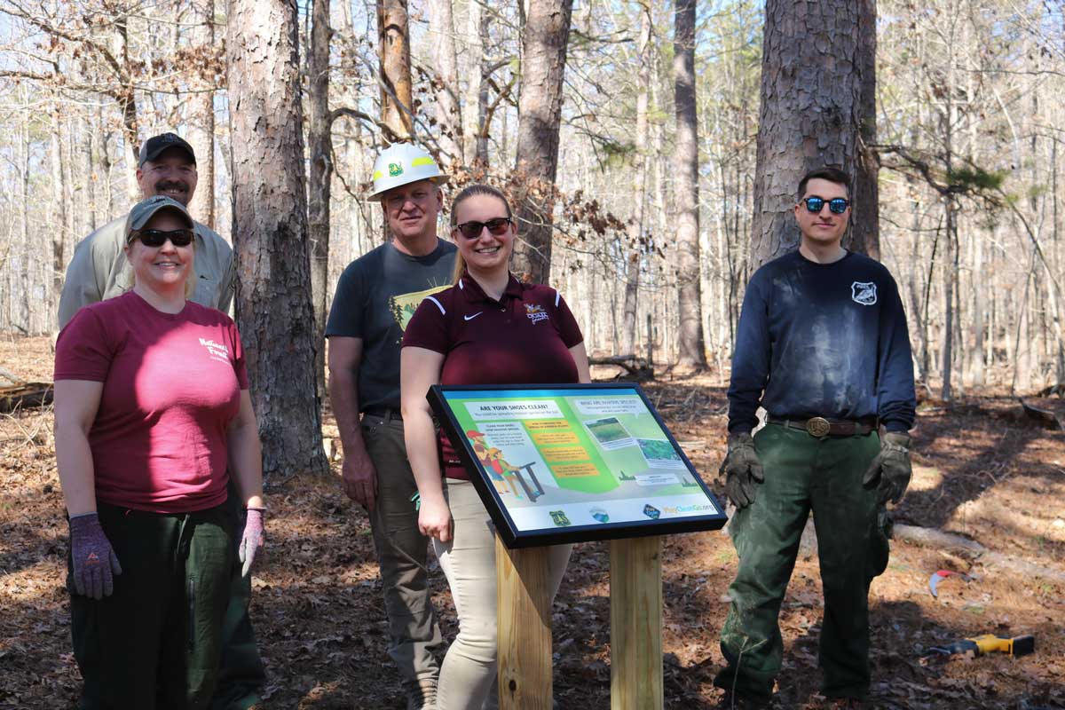 five people wearing casual and business casual clothes in a forest, standing behind a trail sign